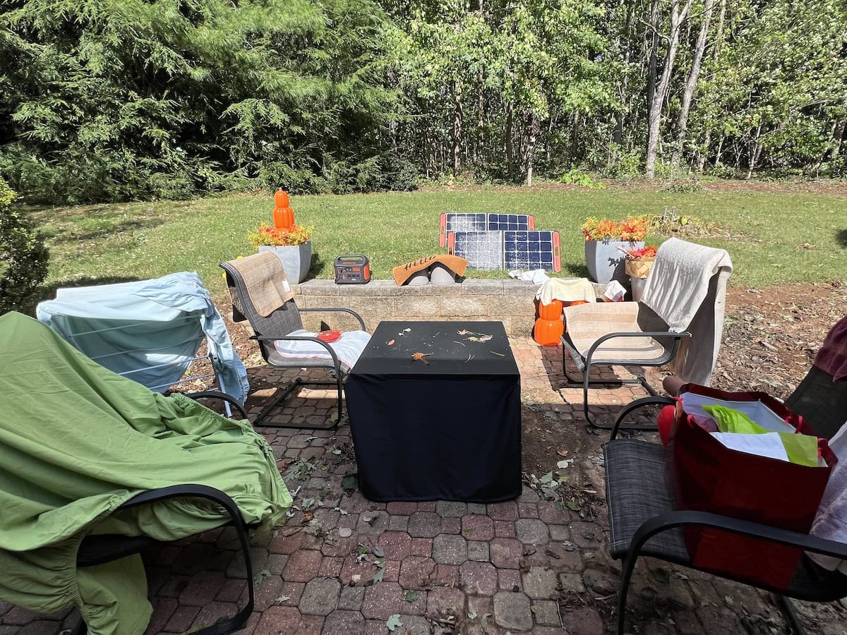 A patio with four chairs surrounding a small table, topped with leaves. Solar panels, plants, and orange structures are in the background, against a backdrop of trees.