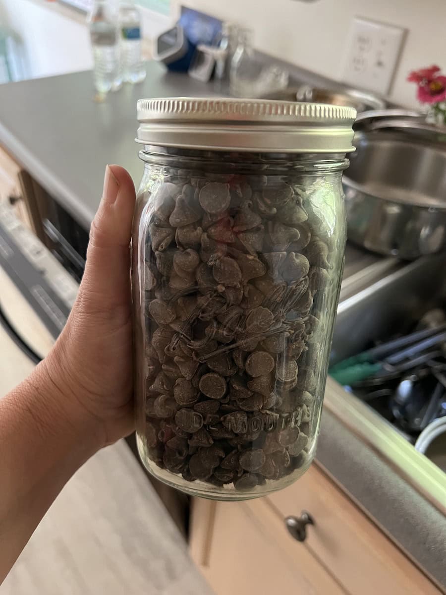 Hand holding a mason jar filled with chocolate chips over a kitchen counter with dishes and water bottles in the background.