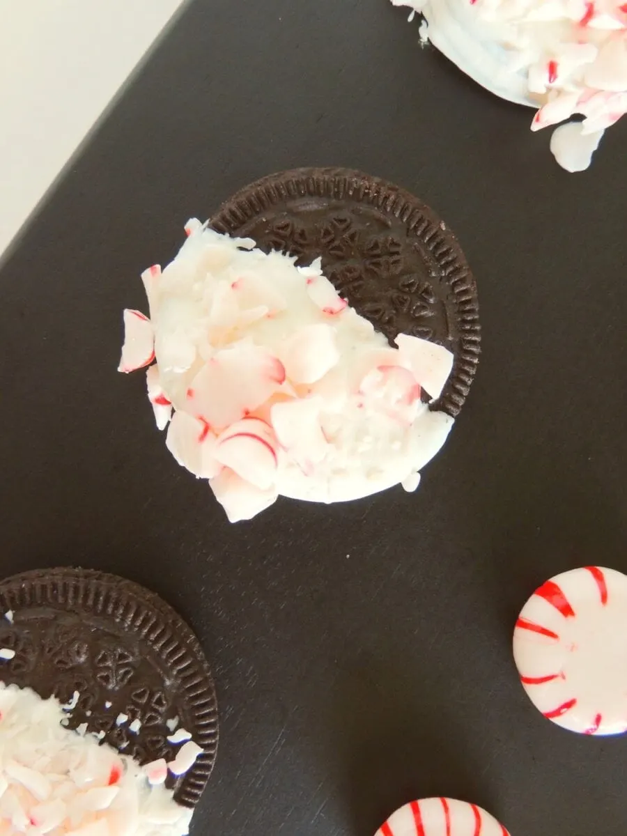 Chocolate sandwich cookie dipped in white icing and topped with crushed peppermint pieces, displayed on a dark surface.