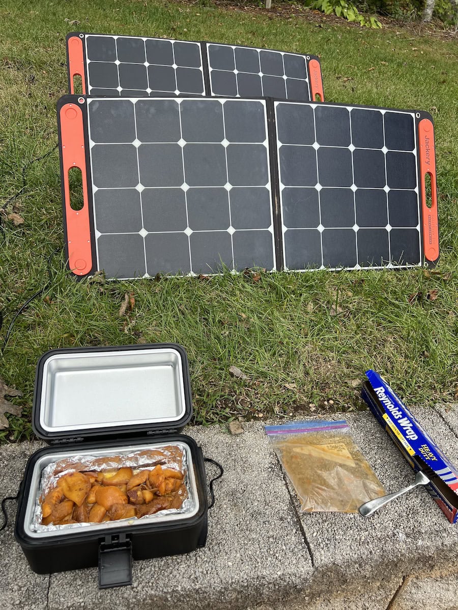 Solar panels on grass with a portable oven cooking food in foil, next to a plastic bag with spices and a box of plastic wrap.