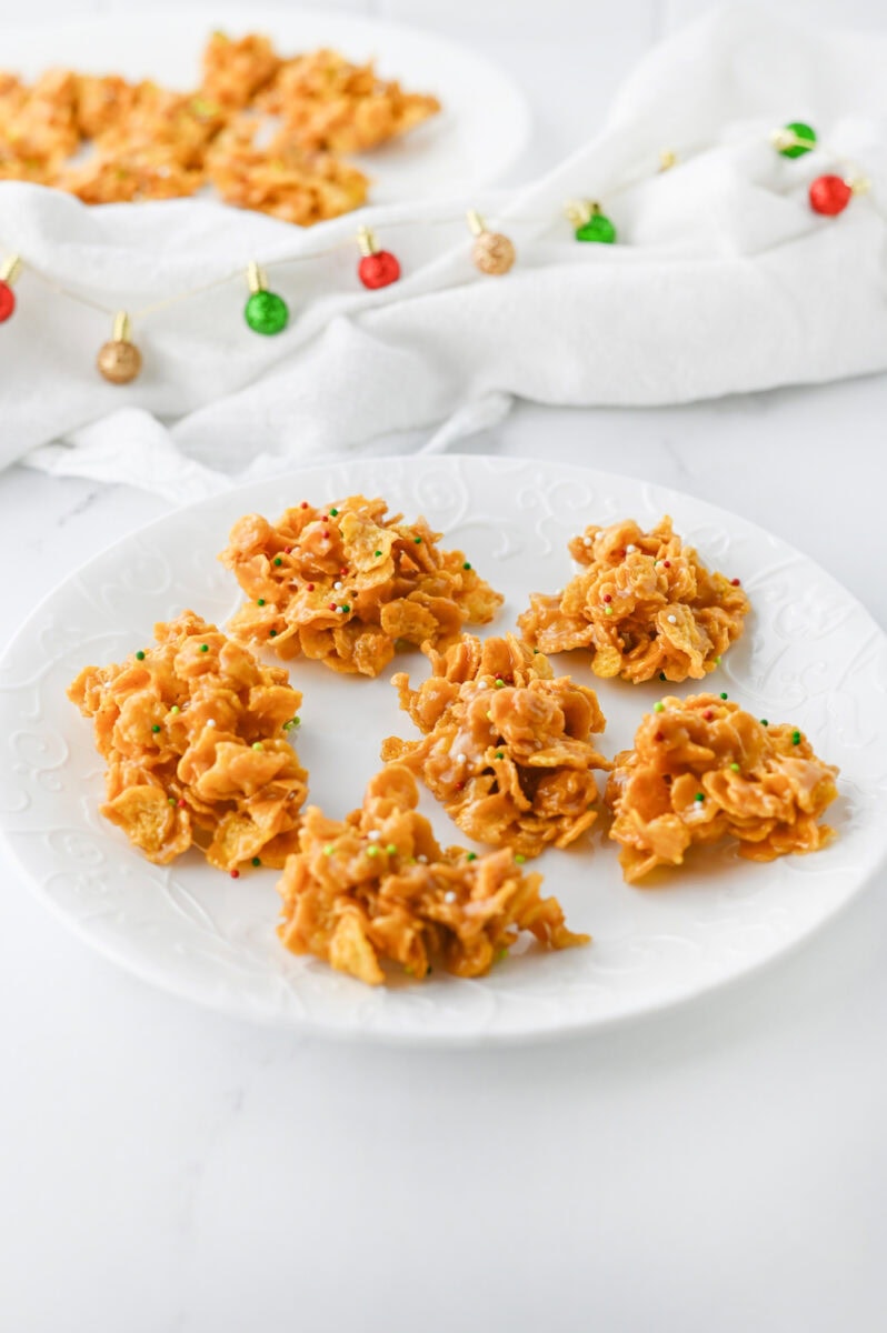 Six cornflake cookies on a white plate with a festive garland in the background.