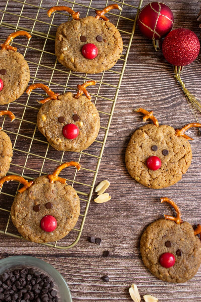 Rudolph-themed cookies on a cooling rack and table, decorated with pretzel antlers, chocolate chip eyes, and red candy noses, surrounded by Christmas ornaments and baking ingredients.