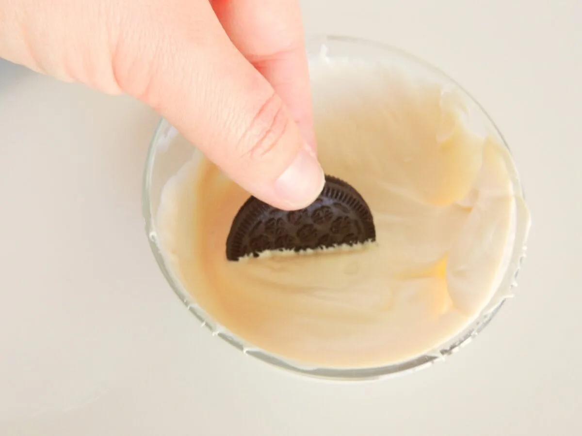 A hand is dipping a chocolate cookie into a bowl of creamy white frosting.