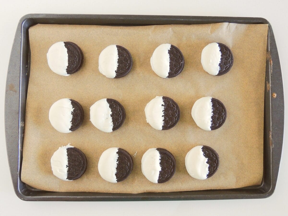 Twelve chocolate sandwich cookies partially dipped in white chocolate on a parchment-lined baking tray.