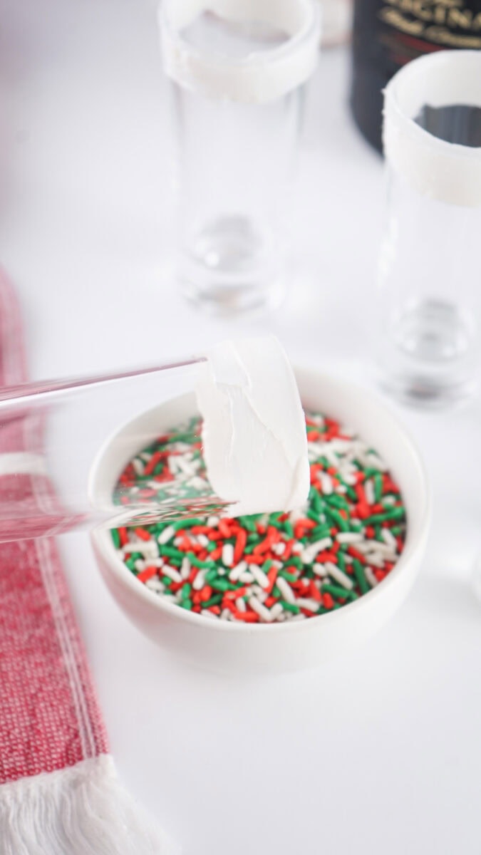 A glass rim with frosting being dipped into a bowl of red, green, and white sprinkles on a white surface.