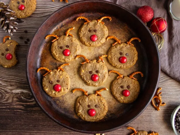 Cookies decorated to resemble reindeer, with pretzel antlers, red candy noses, and chocolate chip eyes, arranged on a round pan.