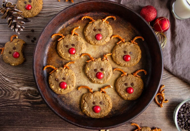 Cookies decorated to resemble reindeer, with pretzel antlers, red candy noses, and chocolate chip eyes, arranged on a round pan.