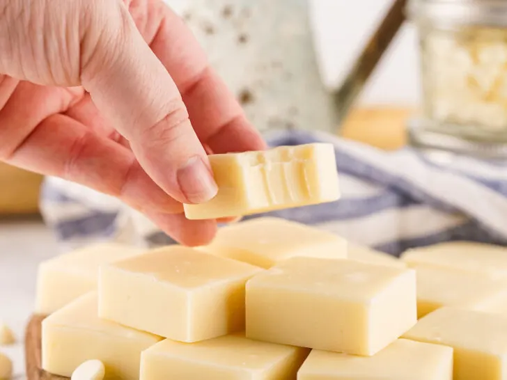 A hand holds a piece of white chocolate with bite marks, hovering above a table laden with even more creamy delights, reminiscent of rich vanilla fudge.