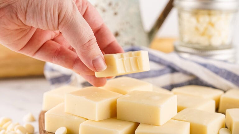 A hand holds a piece of white chocolate with bite marks, hovering above a table laden with even more creamy delights, reminiscent of rich vanilla fudge.