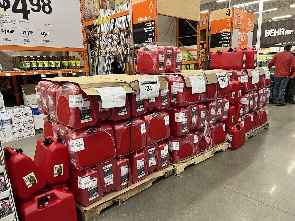 A display of red gas cans on pallets in a hardware store aisle, with price tags visible. Multiple rows and stacks are organized neatly. Shoppers are seen in the background.