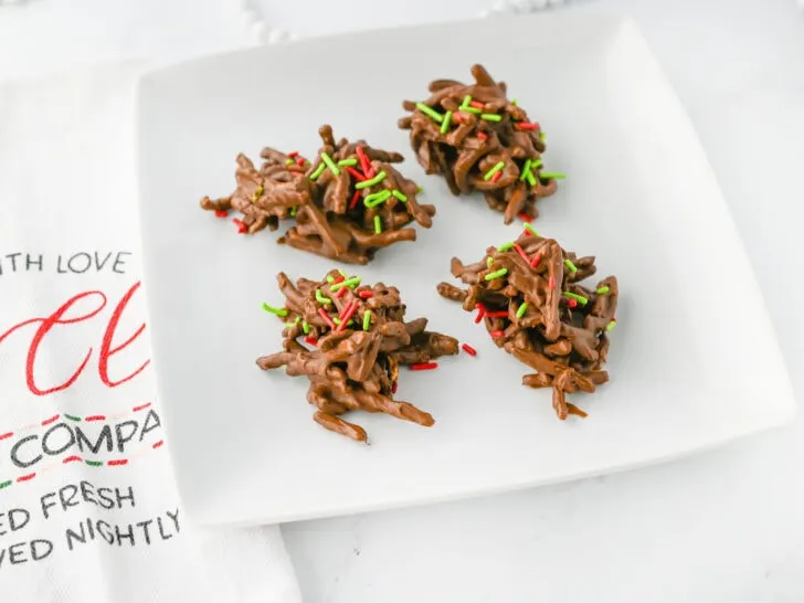 Four chocolate haystack cookies with red and green sprinkles on a white plate.