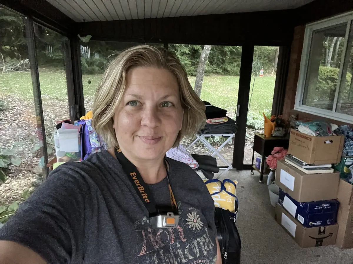 A person with short blonde hair is standing on a porch surrounded by boxes and clutter, smiling at the camera. Large windows reveal a grassy yard outside.