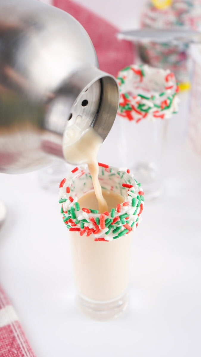 A cocktail shaker pours a creamy drink into a shot glass with a festive red and green sugar rim on a white surface.