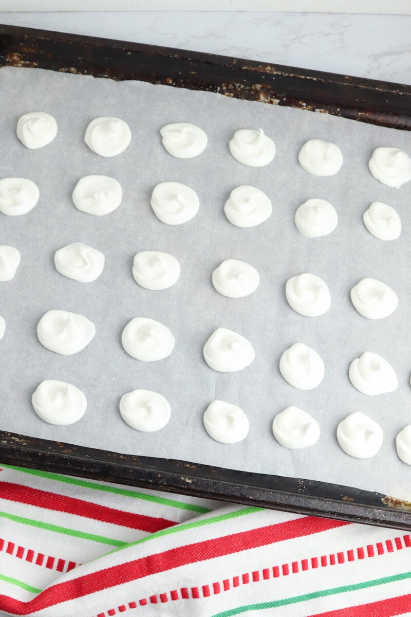 A baking tray lined with parchment paper holds rows of evenly spaced, small white meringue dollops. A striped red, green, and white towel is partially visible underneath the tray.