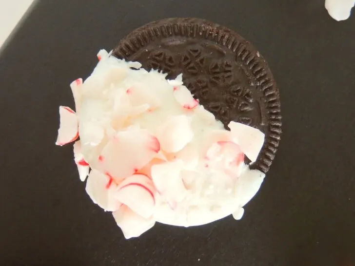 A chocolate sandwich cookie is partially dipped in white icing and covered with small peppermint candy pieces on a dark surface.