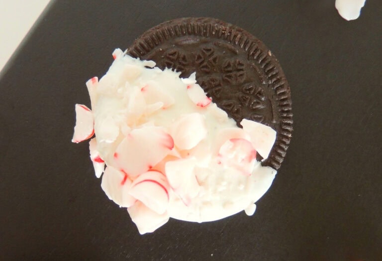 A chocolate sandwich cookie is partially dipped in white icing and covered with small peppermint candy pieces on a dark surface.