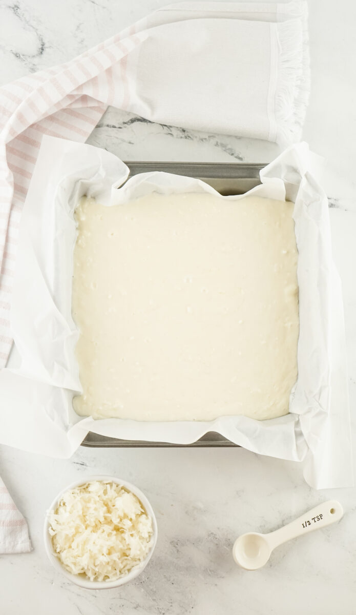 Square baking pan lined with parchment paper filled with white batter. Nearby are a striped cloth, coconut flakes in a bowl, and a measuring spoon.