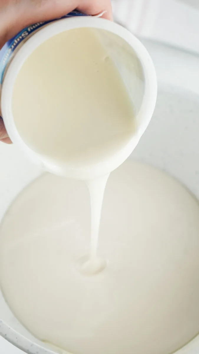 A hand pours a thick, creamy liquid from a white container into a bowl.