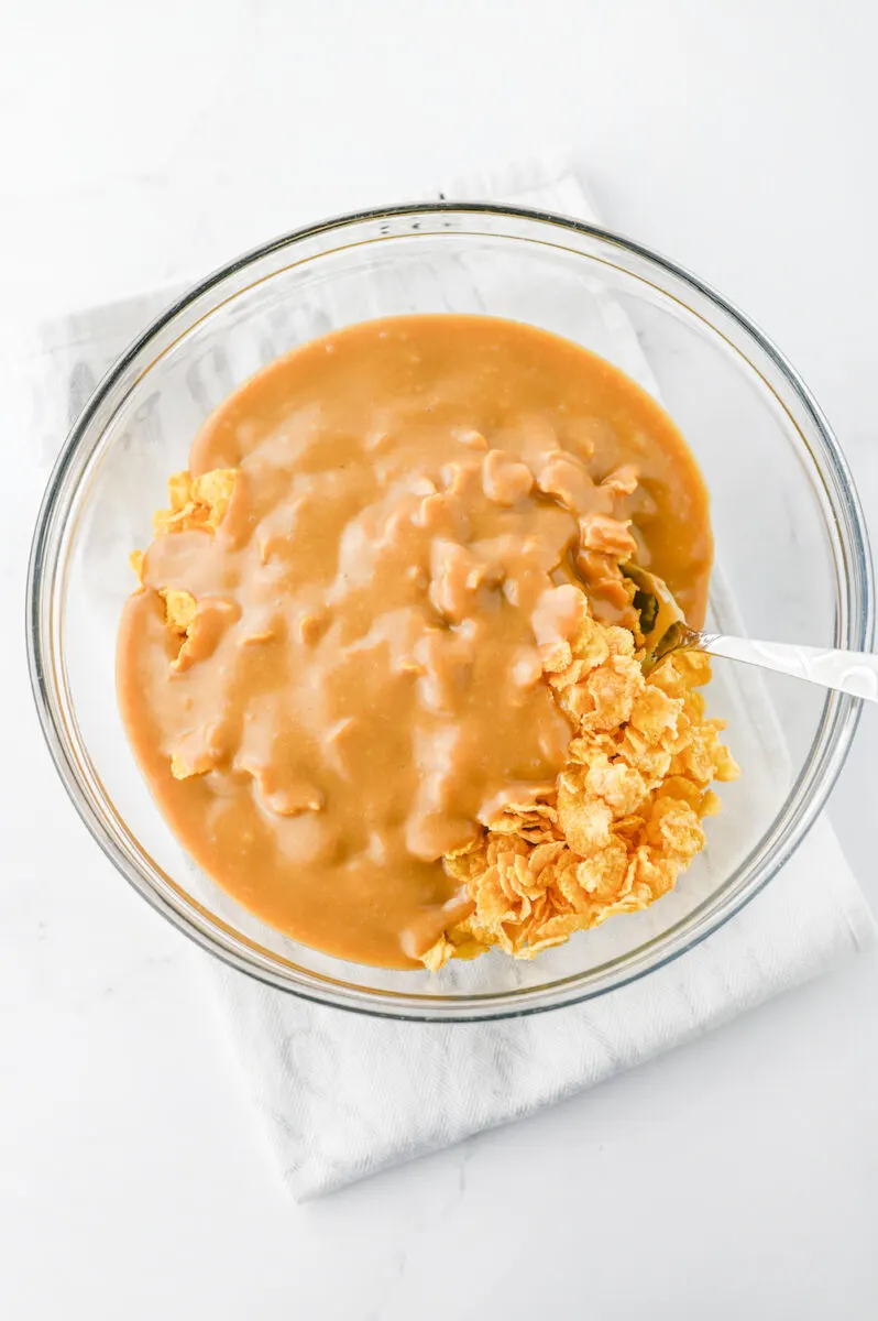 A clear glass bowl filled with cornflakes being mixed with a creamy, caramel-colored sauce. A silver spoon is visible in the mixture, resting on a white cloth.