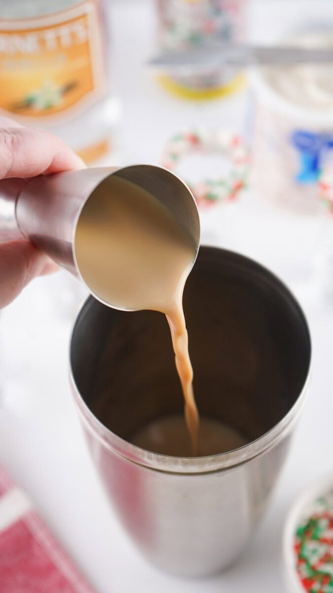 A hand pours Irish cream liqueur from a steel jigger into a cocktail shaker. Various ingredients and decorations are visible in the background.