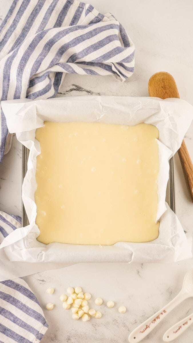 A square baking pan brims with creamy vanilla fudge on parchment paper, surrounded by a wooden spoon, measuring spoons, striped cloth, and white chocolate chips on a marble surface.