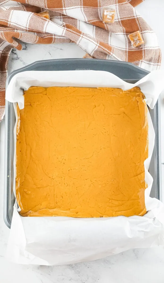 A square pan filled with smooth, orange batter, lined with parchment paper. A checkered cloth is partially visible in the background.