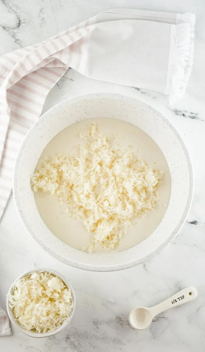 A bowl of shredded coconut in creamy liquid is placed on a marble surface, accompanied by a teaspoon and a cloth on the side.