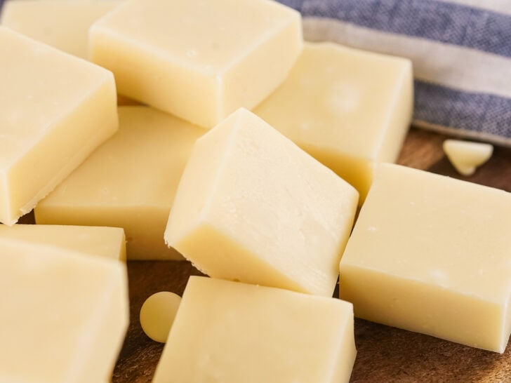 Several blocks of vanilla fudge and a few chocolate chips are arranged on a wooden surface with a striped cloth nearby.