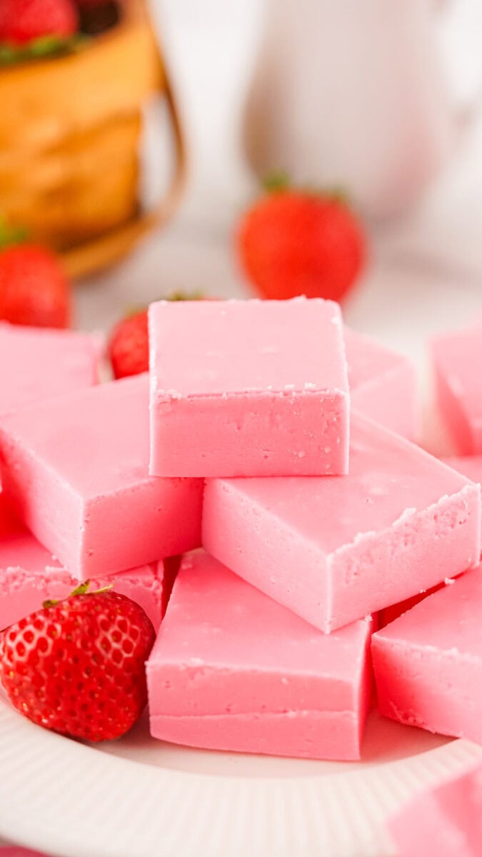 Pink strawberry fudge squares stacked on a plate with whole strawberries and a wicker basket nearby.