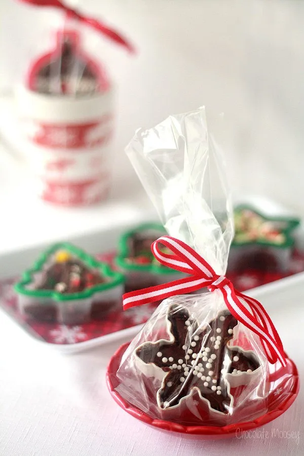 Chocolate snowflake cookies with white sprinkles sit in a clear bag tied with a red ribbon. Holiday-themed cups and green tree-shaped cookies complete the festive scene, perfect for pairing with homemade fudge recipes on a white surface.