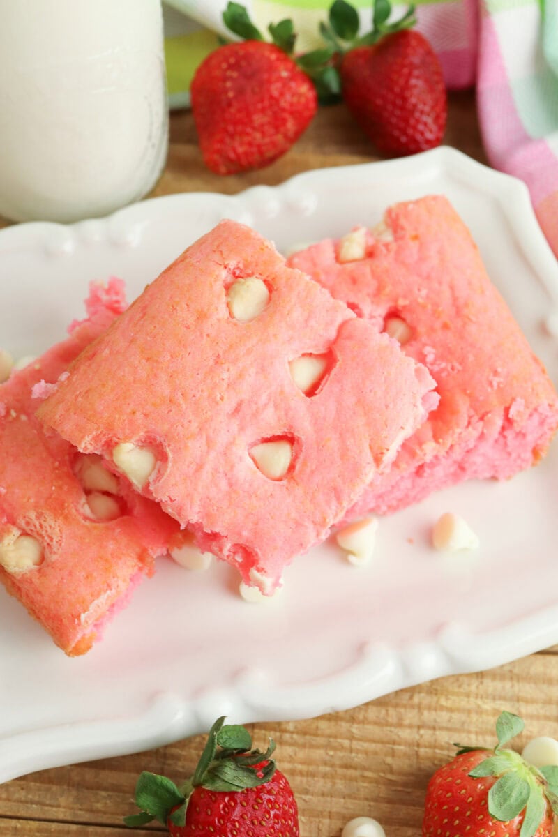 Three square pink cake pieces with white chocolate chips on a white plate, surrounded by fresh strawberries.