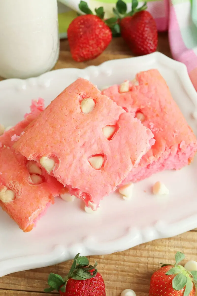 Three square pink cake pieces with white chocolate chips on a white plate, surrounded by fresh strawberries.