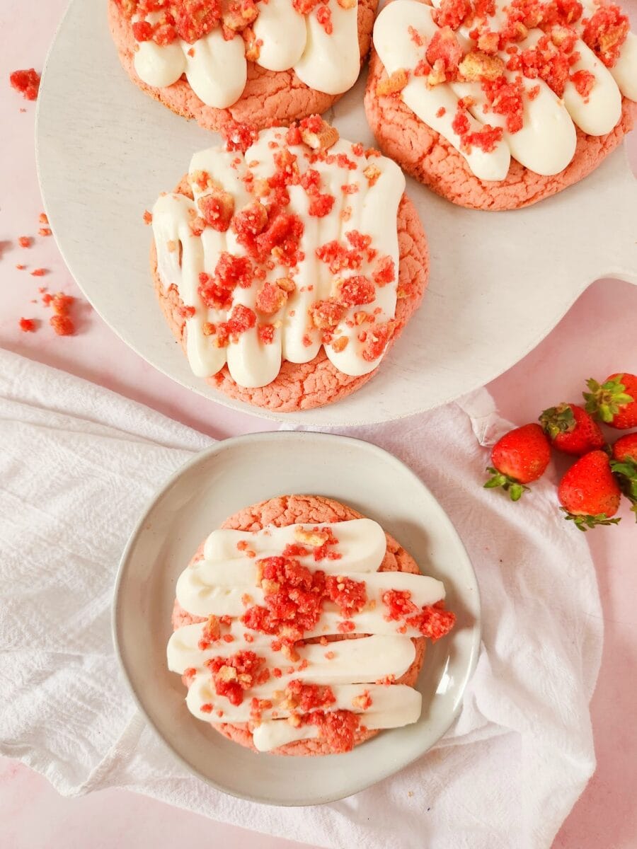 Two pink cookies with white frosting and red crumbles on a white plate and pedestal. Fresh strawberries are scattered nearby on a white cloth.