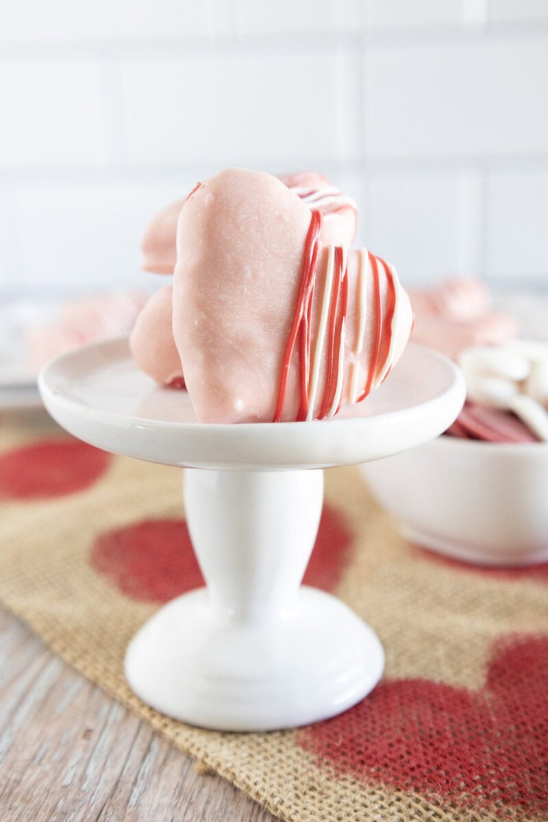 Heart-shaped pink cookies with red stripes on a white pedestal, set on a textured surface with a blurred background.