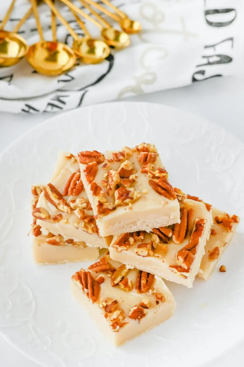 A stack of pecan pie fudge pieces, rich and creamy, topped with chopped pecans on a white plate. Gold measuring spoons and a patterned cloth provide an elegant backdrop.