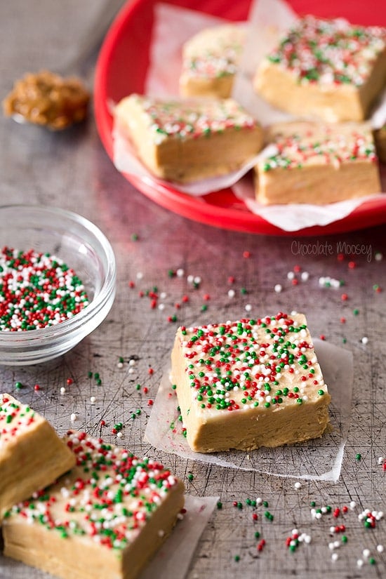 Squares of peanut butter fudge from homemade fudge recipes are topped with red, green, and white sprinkles on a tabletop and a red plate. A glass bowl with extra sprinkles is nearby.