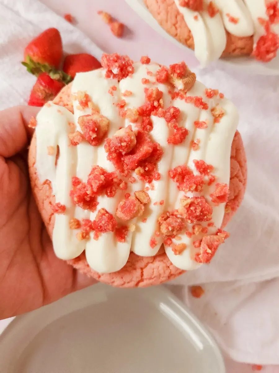 Hand holding a pink cookie topped with white icing and pink crumbles, on a light background with a couple of strawberries nearby.