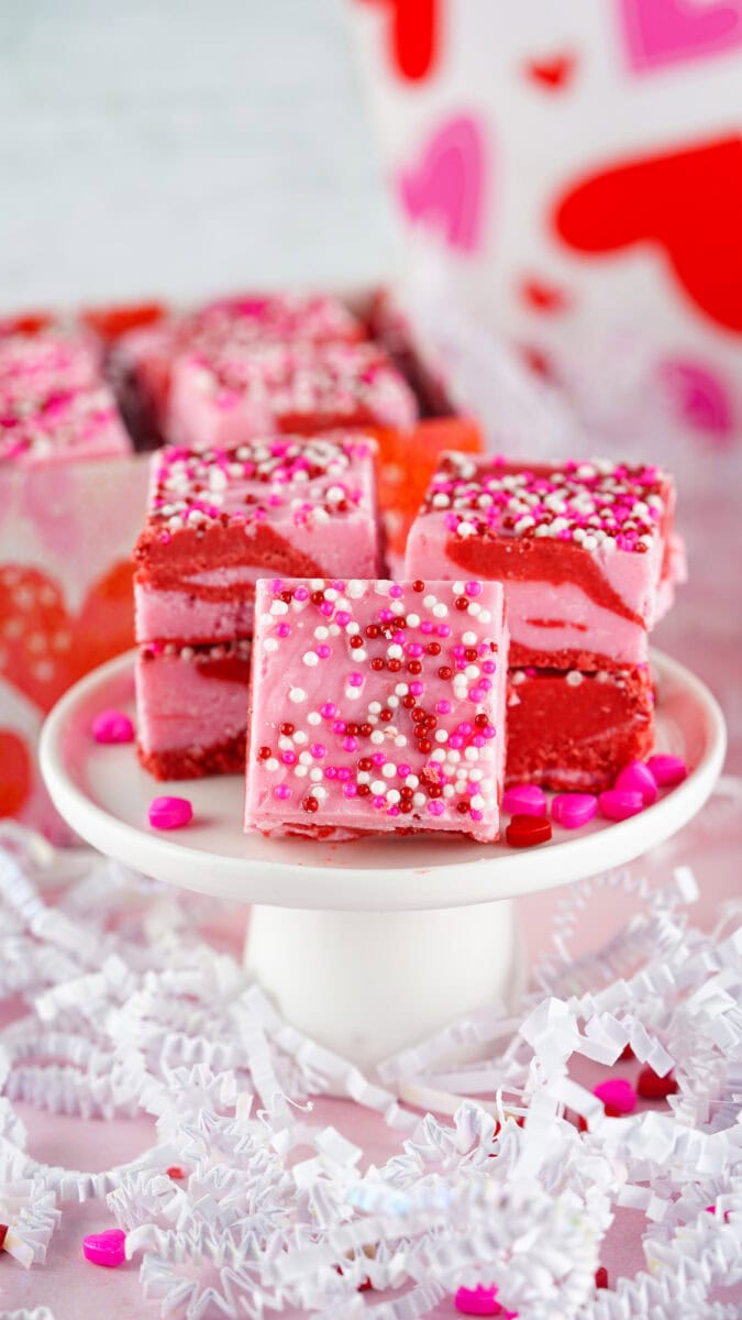 Close-up of pink frosted cake squares with colorful sprinkles on a white pedestal, surrounded by decorative paper shreds and candy.