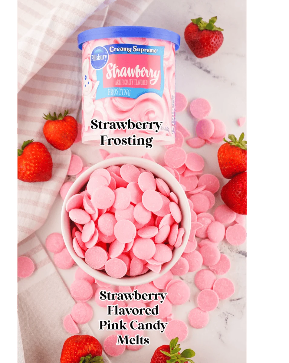 Container of strawberry frosting and a bowl of pink candy melts on a marble surface, surrounded by fresh strawberries.