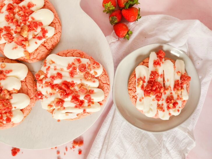 Strawberry cookies with white icing and red crumbles on a plate, surrounded by whole strawberries on a pink surface.