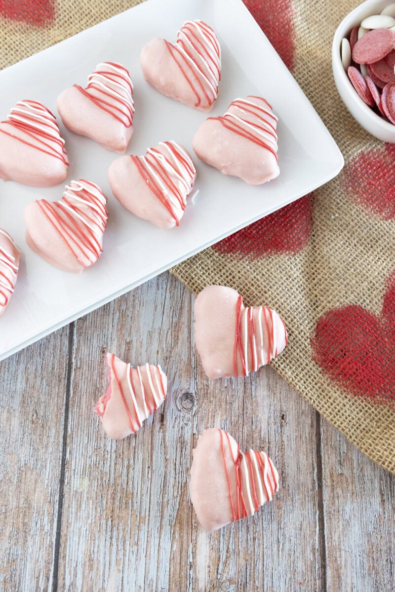 Heart-shaped cookies with pink frosting and red drizzle on a white plate, on a rustic wooden table with a burlap heart-patterned cloth. A bowl of red chocolate pieces nearby.