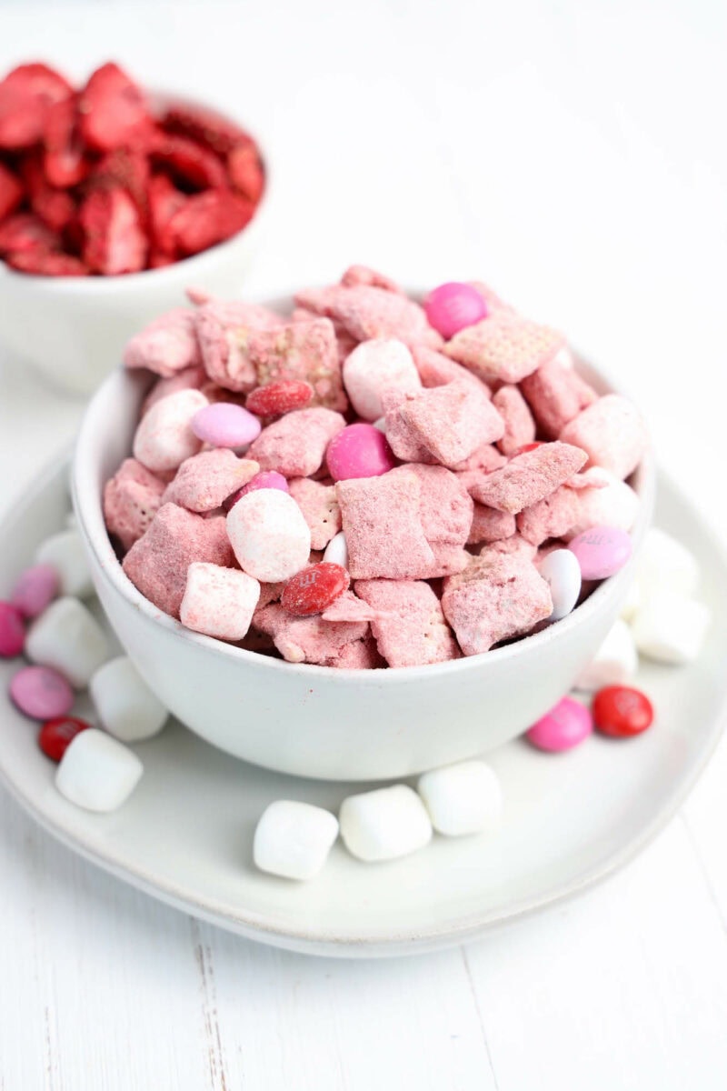 A bowl of pink and white snack mix with marshmallows and candies. Another bowl with red snacks is partially visible in the background.