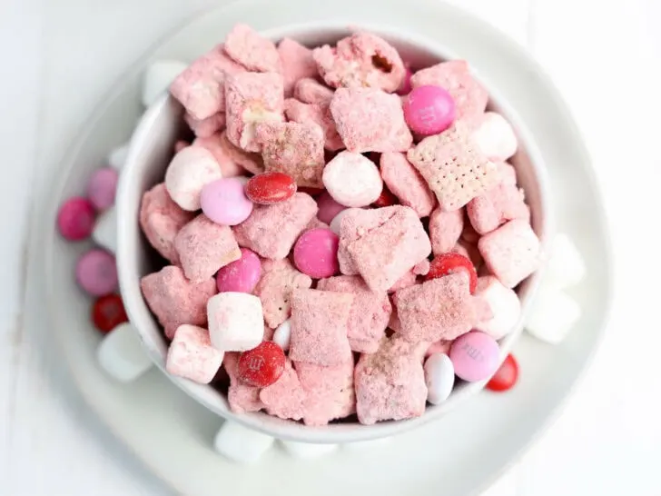 A white bowl filled with pink and white cereal, candy-coated chocolates, and marshmallows on a white plate.