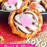 Close-up of pretzel treats adorned with chocolate, a pink heart candy, and "Hugs & Kisses Pretzel Treats" text at the bottom. The heartwarming display sits on a red textured surface, embodying the message of hugs and kisses.