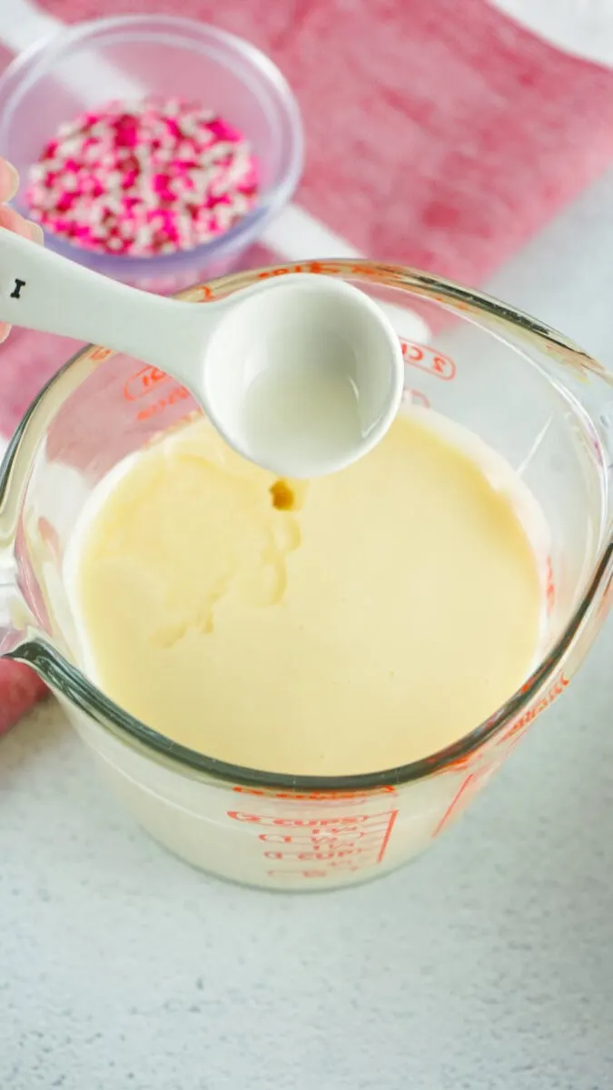 A measuring cup with vanilla pudding is being mixed, with sprinkles nearby on a textured countertop.