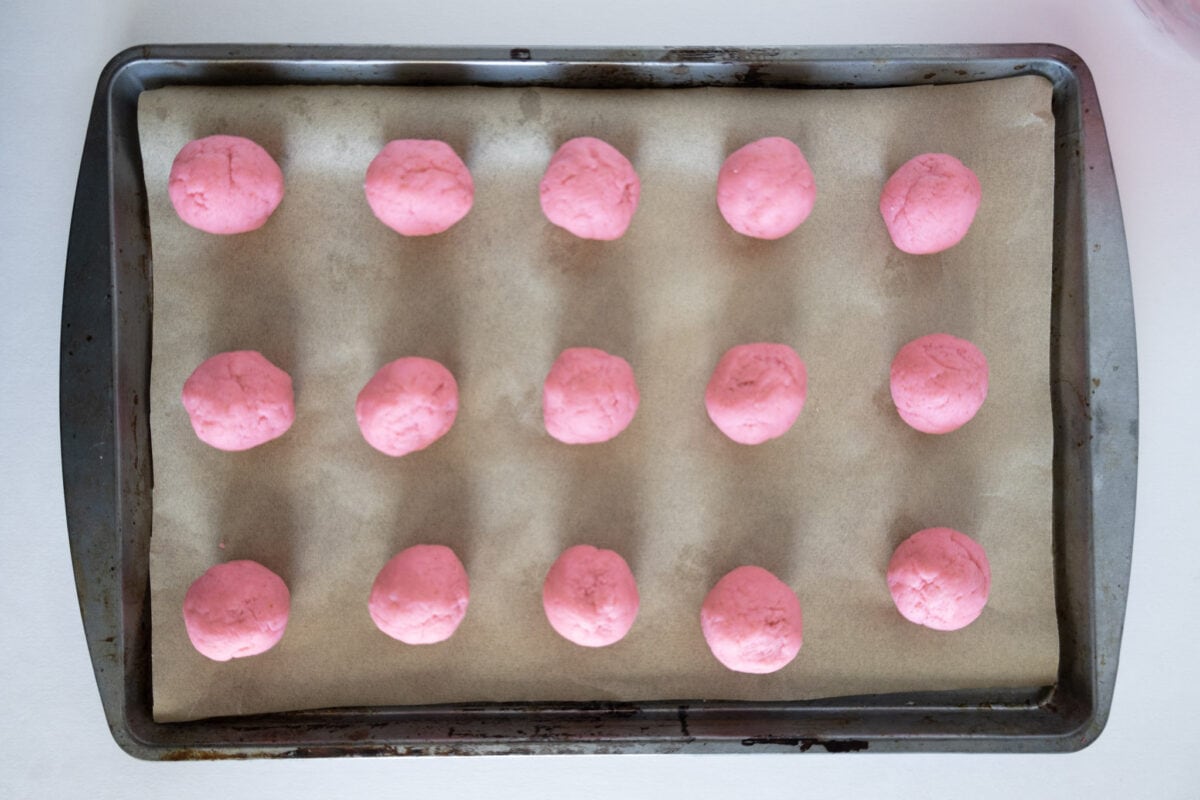 A baking tray with 16 evenly spaced pink dough balls on parchment paper.