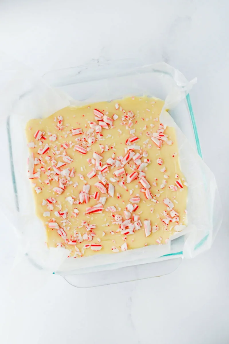 Glass dish with white chocolate fudge topped with crushed candy canes on a parchment-lined surface.