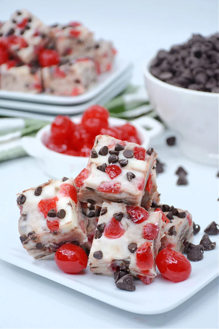 Slices of chocolate chip cherry fudge, crafted from homemade fudge recipes, rest on a white plate surrounded by whole cherries and extra chocolate chips.