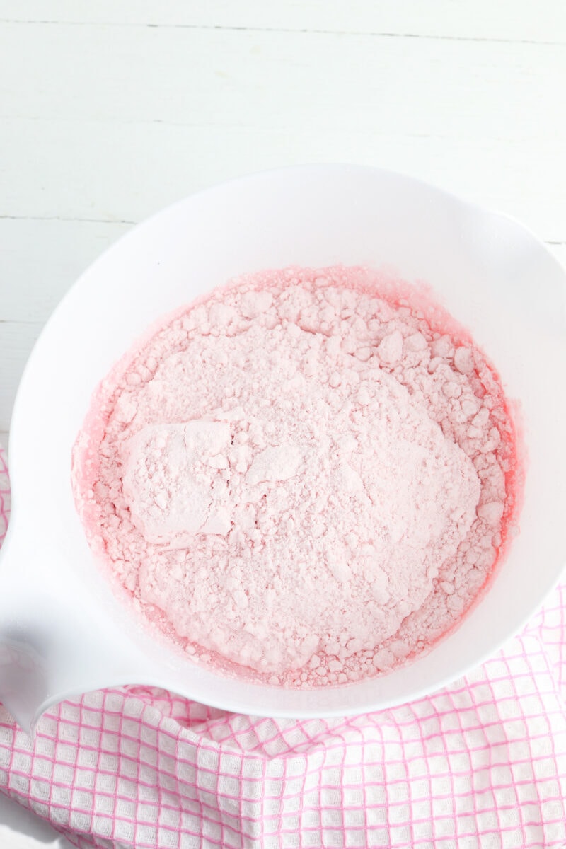 A white mixing bowl filled with pink powder sits on a white surface beside a pink checkered cloth.