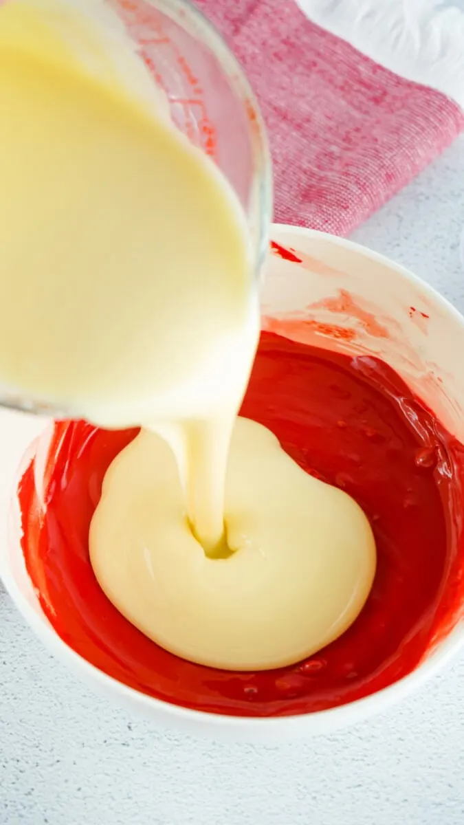 Cream-colored liquid being poured into a bowl containing red liquid. A red cloth is visible in the background.